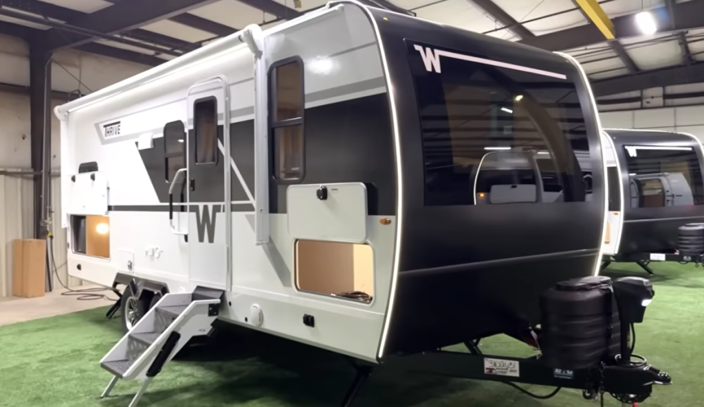 Winnebago Thrive travel trailer displayed in an indoor showroom, featuring a sleek black front cap with panoramic window, LED accent lighting, dual entry steps, and exterior storage compartments illuminated for a premium modern look.