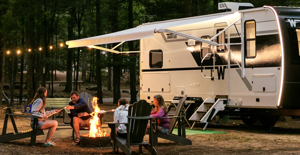 Family sitting around a campfire roasting marshmallows beside a Winnebago Thrive travel trailer with LED awning lights extended, showcasing the modern black-and-white exterior and cozy campsite atmosphere in a wooded setting.
