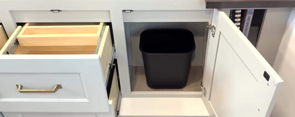 Brinkley Model G Series luxury RV interior featuring a hidden trash bin storage cabinet beside deep drawers with gold hardware and soft-close functionality, showcasing smart residential-style kitchen organization.