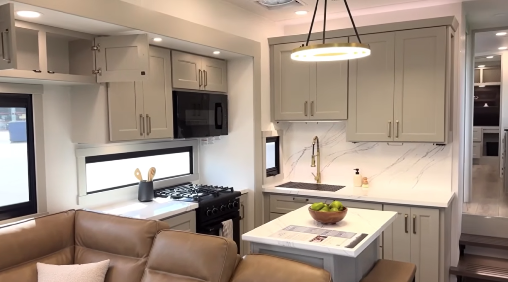 Brinkley Model G Series RV kitchen featuring modern cabinetry, solid-surface countertops, farmhouse sink with gold faucet, built-in microwave and gas stove, under-cabinet lighting, and a central island for dining and prep space.