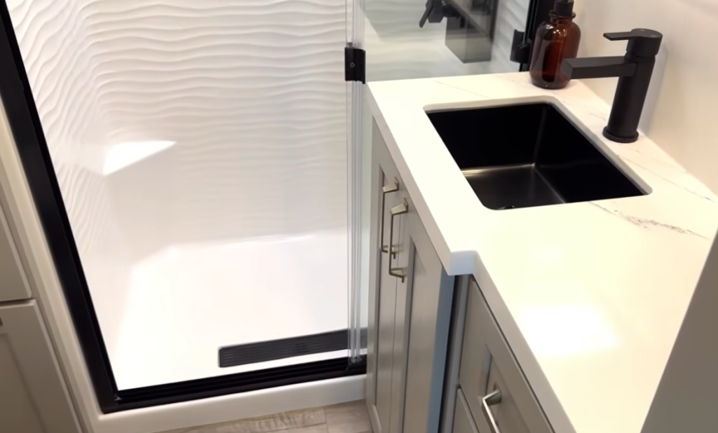 Brinkley Model G Series RV bathroom with solid-surface vanity top, modern black faucet and sink, and glass-enclosed shower featuring a textured wall design for a spa-like, residential finish.