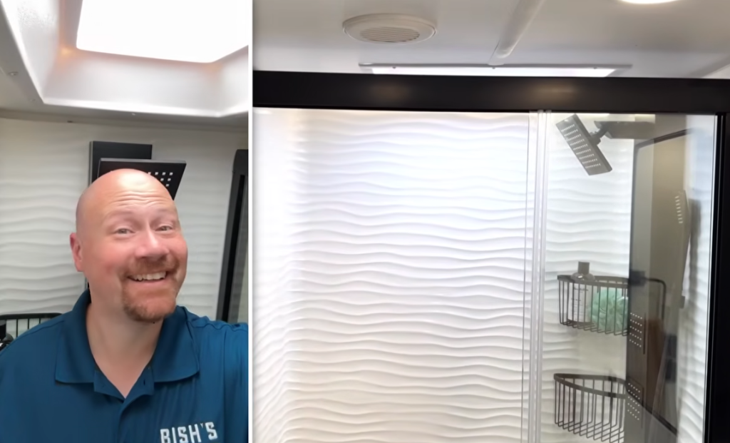 Brinkley Model G Series RV bathroom featuring a large glass-enclosed shower with textured wave wall, rainfall showerhead, and storage baskets, shown by a Bish’s RV representative during a walkthrough.