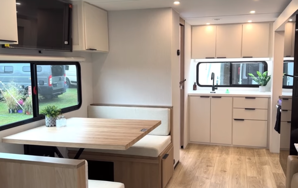 2026 Winnebago Thrive 22MLS interior view featuring a bright kitchen with white cabinetry, black hardware, under-window lighting, and a spacious dinette with wood table and large panoramic windows.