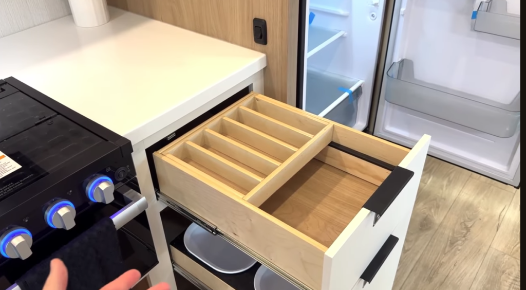 2026 Winnebago Thrive 22MLS kitchen featuring organized pull-out drawers with utensil dividers beside a black three-burner stove and open refrigerator, highlighting smart storage design.