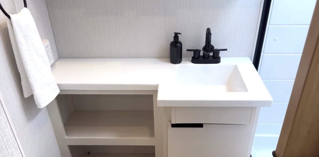 2026 Winnebago Thrive 22MLS bathroom vanity with seamless white countertop, modern black faucet, open shelving, and towel ring for a clean, functional design.