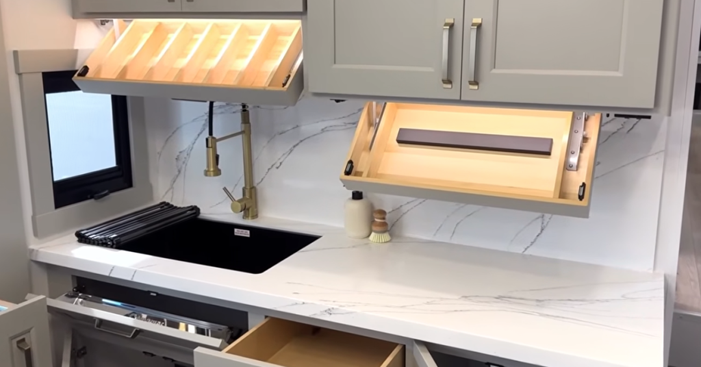 “Brinkley Model G Series RV kitchen showcasing illuminated overhead cabinets with built-in spice rack and dish storage, gold pull-down faucet, deep farmhouse sink, marble-look countertop, and luxury under-cabinet lighting.