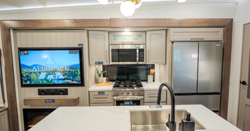 Alliance Paradigm fifth wheel kitchen with island sink, residential refrigerator, microwave, gas range, and entertainment center featuring a large TV and electric fireplace in a modern, bright interior.