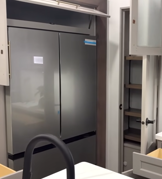 Alliance Paradigm fifth wheel kitchen showing stainless steel residential refrigerator, open pantry with adjustable shelving, and upper storage cabinet above the fridge for added organization.