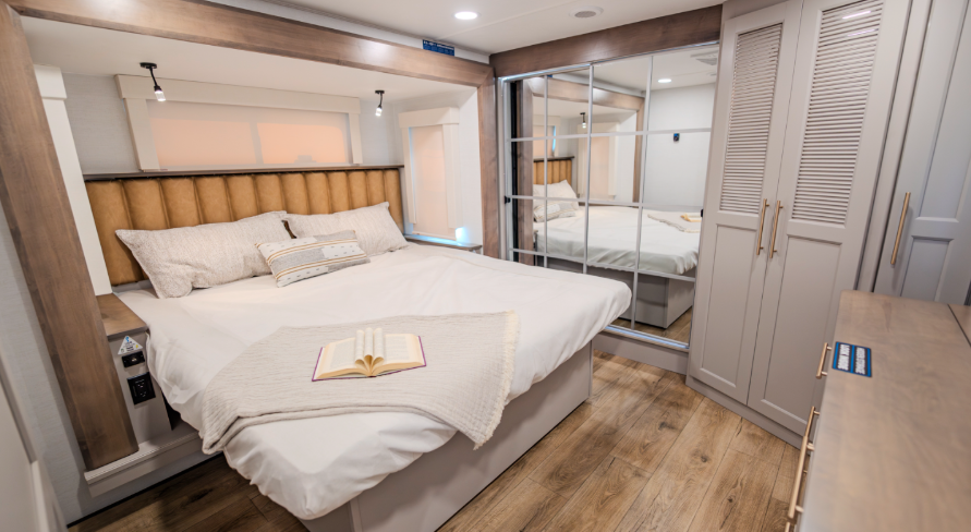 Alliance Paradigm fifth wheel bedroom featuring a king bed with padded headboard, mirrored wardrobe closets, wood-look flooring, and soft ambient lighting for a residential feel.