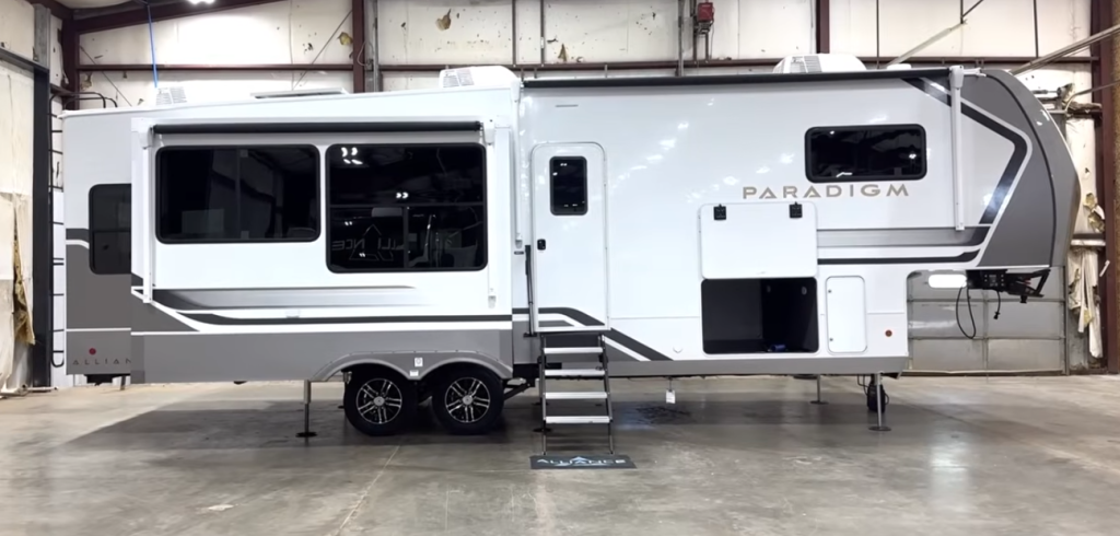 2026 Alliance Paradigm fifth wheel exterior side profile with extended slide-outs, alloy wheels, and dual-entry storage compartments inside a display facility