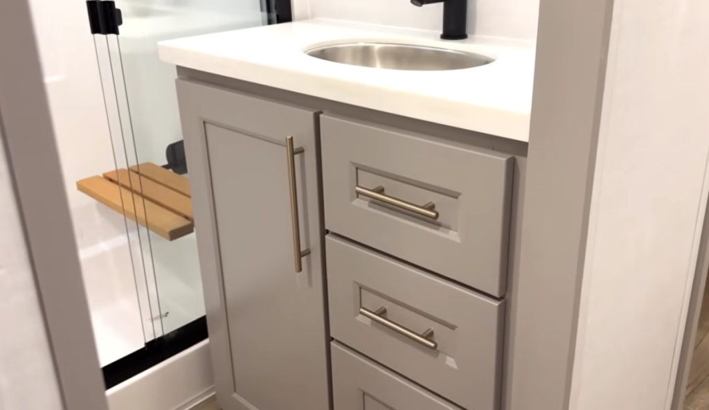 Alliance Paradigm fifth wheel bathroom vanity with solid-surface countertop, stainless steel sink, matte black faucet, and storage drawers beside the glass shower with teak bench.