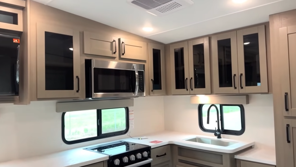 2026 Grand Design Reflection 150 270BN kitchen with U-shaped counter, stainless steel microwave, gas range, black faucet, and overhead glass-front cabinetry in a modern neutral finish.