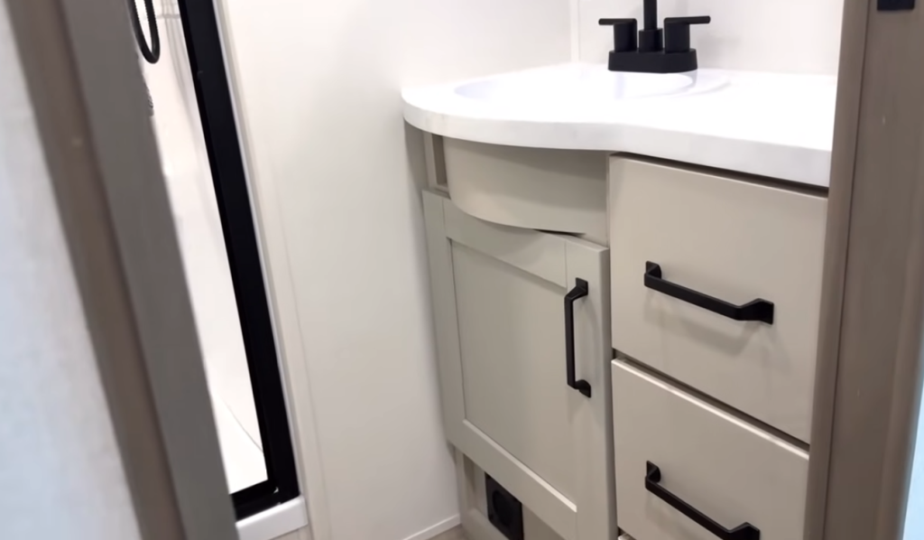2026 Grand Design Reflection 150 270BN bathroom vanity with curved countertop, black faucet, and light cabinetry offering modern style and practical storage space.