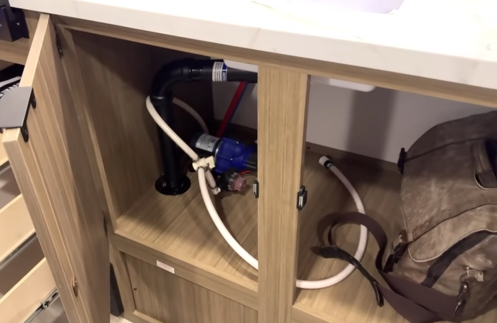 Under-sink storage area in the Forest River Wildwood FSX showing accessible plumbing, water pump, and PEX lines beneath the kitchen countertop.