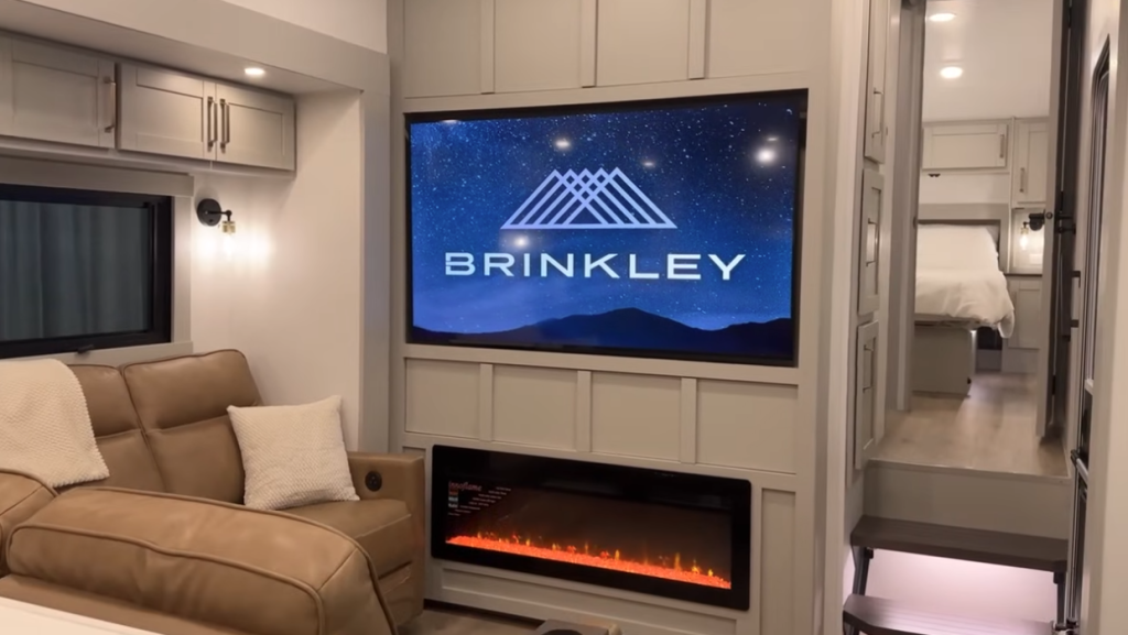 Brinkley fifth wheel RV living area featuring a large HDTV with the Brinkley logo, modern electric fireplace, tan leather recliners, and a bright open layout leading to the master bedroom.
