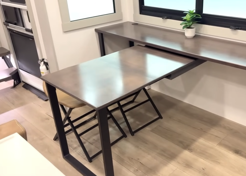 Brinkley Model Z fifth wheel multifunctional desk and dining space featuring a pull-out table with modern black metal legs, folding chairs, and a minimalist workspace under large windows.