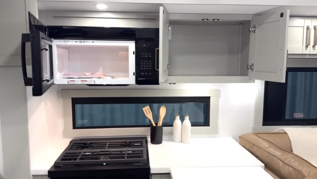 Brinkley Model Z RV kitchen showcasing a built-in black microwave above a three-burner gas stove, bright backsplash window, and overhead cabinetry for efficient storage and modern functionality.