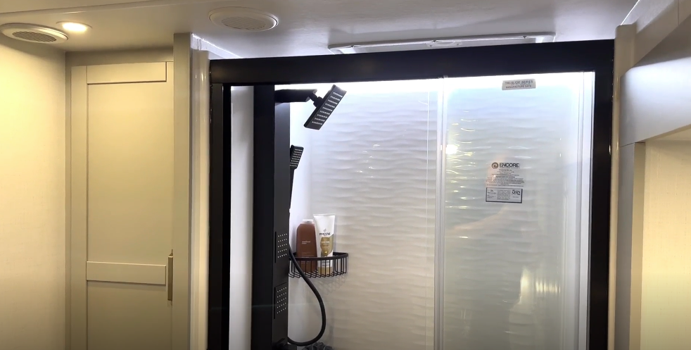 Brinkley Model Z RV bathroom shower with textured white surround, black rainfall shower system, glass sliding doors, and built-in corner shelving for a modern residential spa feel.