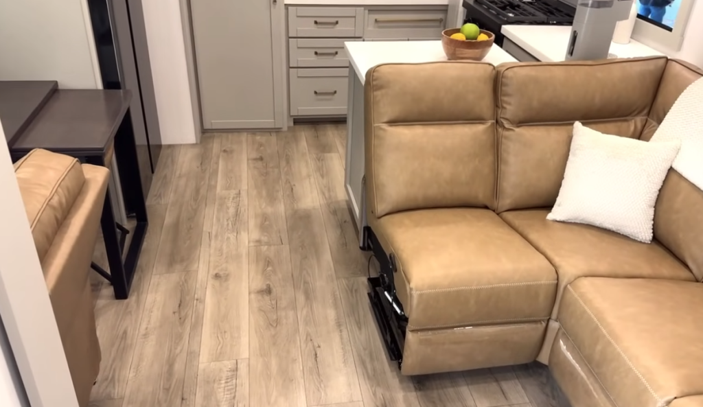Brinkley Model Z living area featuring a tan leather power recliner sectional, light wood plank flooring, and open floor space leading into the modern kitchen with gray cabinetry and island counter.