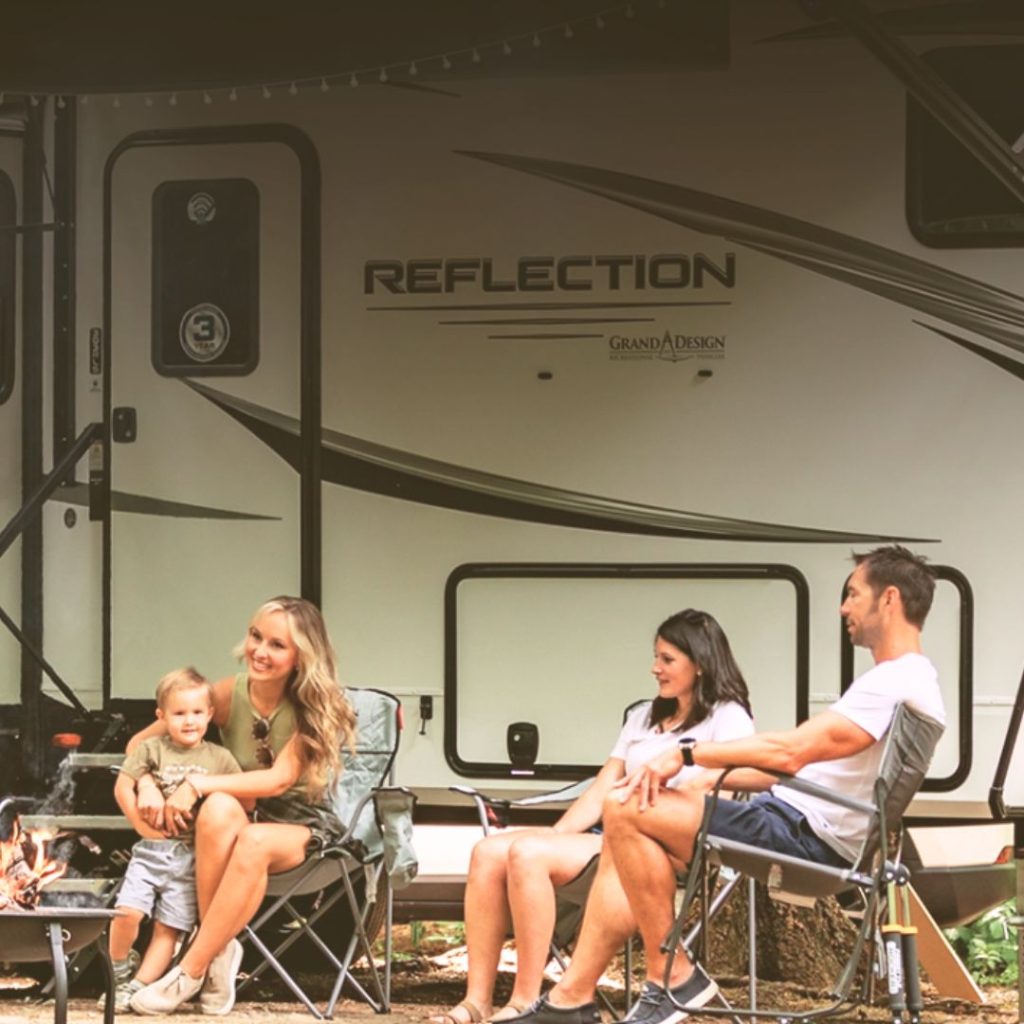 Family relaxing outside a Grand Design Reflection fifth wheel, sitting around a campfire in folding chairs, enjoying a cozy outdoor camping evening.