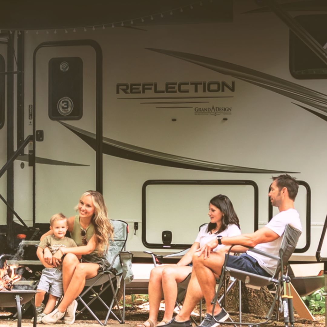 Family relaxing outside a Grand Design Reflection fifth wheel, sitting around a campfire in folding chairs, enjoying a cozy outdoor camping evening.