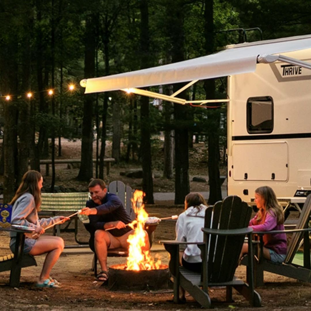 Family camping beside a Winnebago Thrive travel trailer, roasting marshmallows around a campfire under string lights and an extended awning in a wooded campground at dusk.