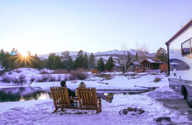 Couple relaxing in wooden chairs beside a snowy lakeside campground with an RV parked nearby, surrounded by mountains at sunset.