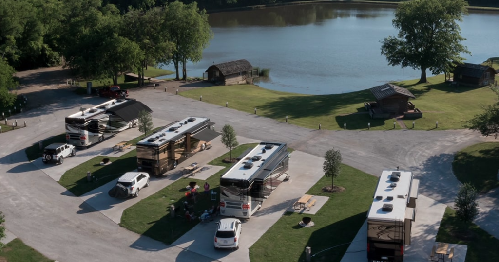 Aerial view of a scenic RV campground beside a lake, with multiple motorhomes parked on paved sites, surrounded by trees, picnic tables, and small wooden cabins near the water.