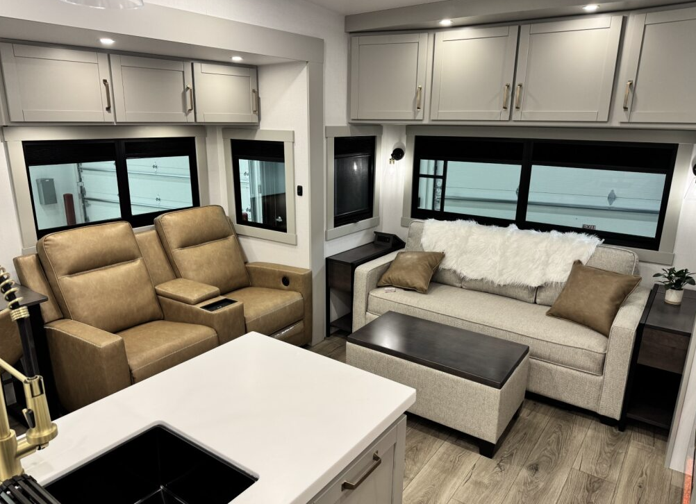 Brinkley Model Z 3600 living room with tan theater seating, tri-fold sofa, overhead storage cabinets, large windows, and a central ottoman for a residential, cozy feel.