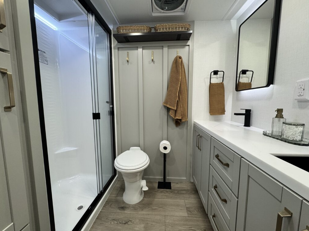 Brinkley Model Z 3600 bathroom featuring a large glass-enclosed shower, porcelain toilet, modern vanity with black fixtures, LED mirror, and soft gray cabinetry with gold hardware.