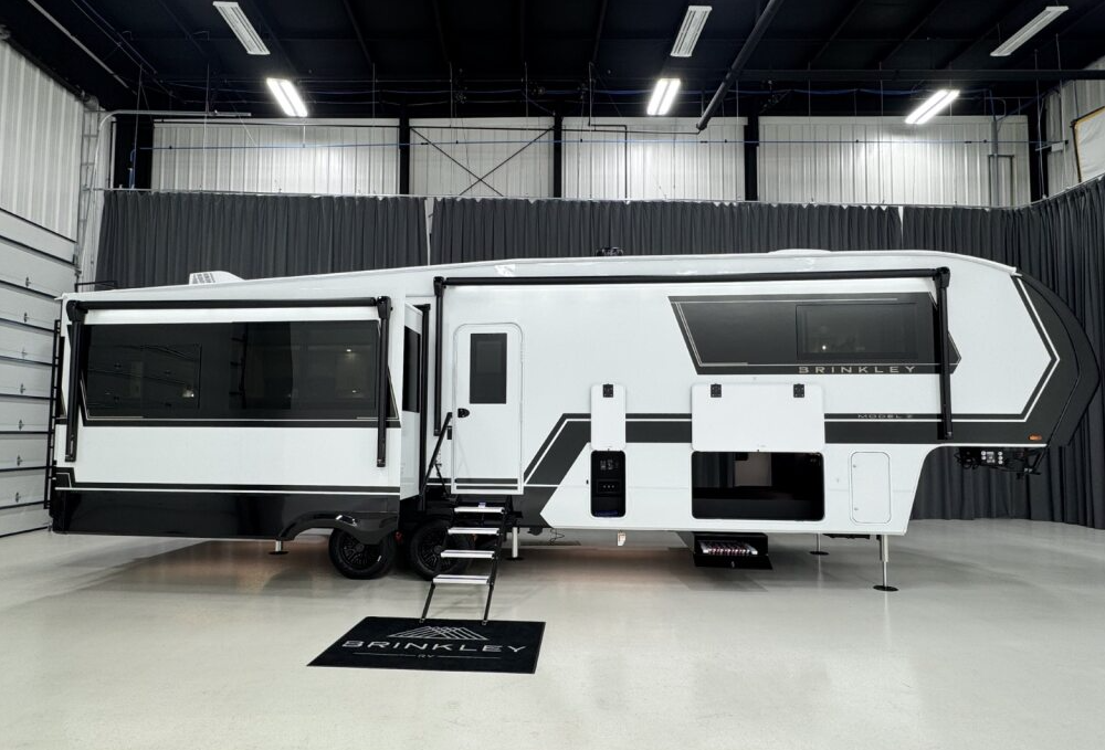 Side profile of a Brinkley Model Z luxury fifth wheel RV shown indoors with slide-outs extended, highlighting its aerodynamic design, black-and-white full-body paint, and premium construction details.