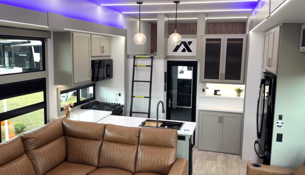 Modern kitchen inside the 2026 Axiom Vendetta 4250 fifth wheel featuring LED accent lighting, a loft access ladder, full-size refrigerator, island sink, stainless gas range, and contemporary gray cabinetry with gold hardware.