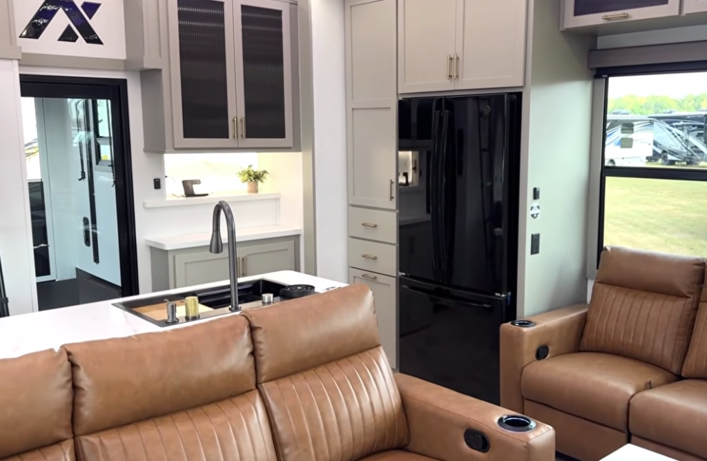 Kitchen and living area inside the 2026 Axiom Vendetta 4250 fifth wheel featuring a modern island sink with high-arc faucet, black residential refrigerator, gray cabinetry with gold hardware, and tan leather recliners.