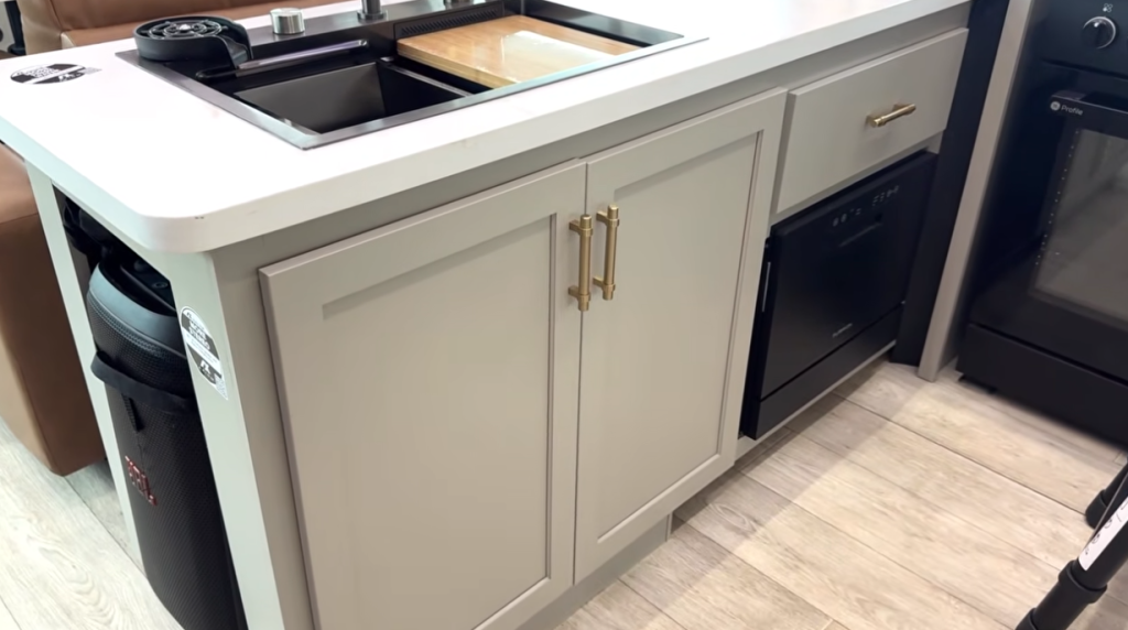 Close-up of the 2026 Axiom Vendetta 4250 kitchen island featuring a deep stainless sink with cover, gold hardware cabinetry, built-in dishwasher, and convenient storage for a portable JBL speaker.