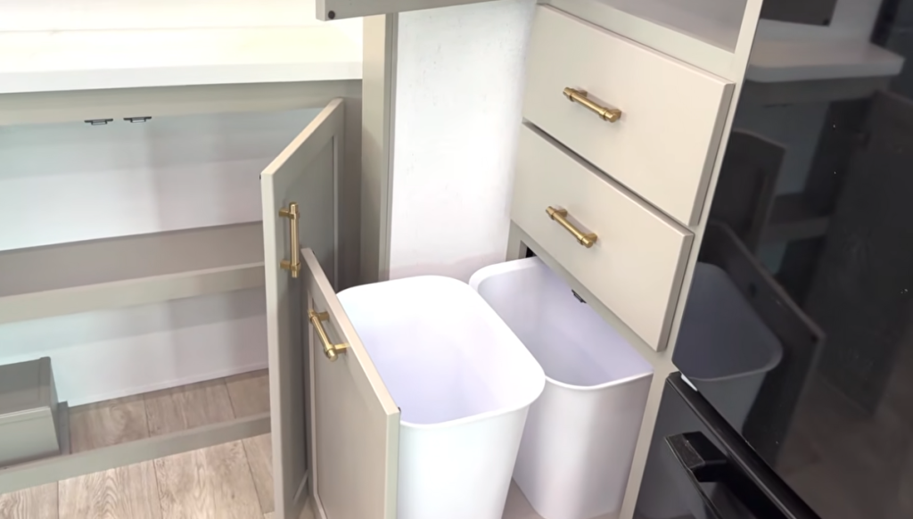 Dual pull-out trash bins built into the kitchen cabinetry of the 2026 Axiom Vendetta 4250 fifth wheel, featuring gold hardware and soft-close drawers for efficient waste and recycling storage.