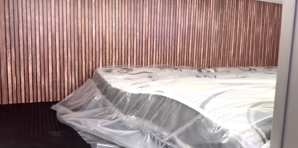 Loft bed area inside the 2026 Axiom Vendetta 4250 fifth wheel featuring a new mattress still wrapped in protective plastic and a modern vertical wood-slat accent wall for added warmth and style.