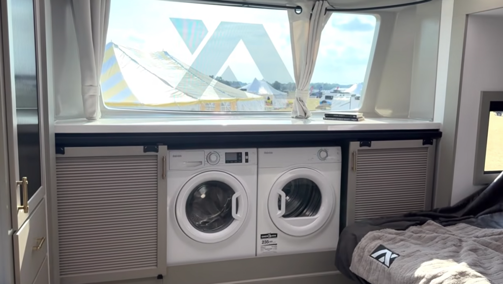 Front bedroom of the 2026 Axiom Vendetta 4250 fifth wheel featuring a panoramic windshield, dual washer and dryer units beneath the window, and modern cabinetry beside a luxury RV king bed.