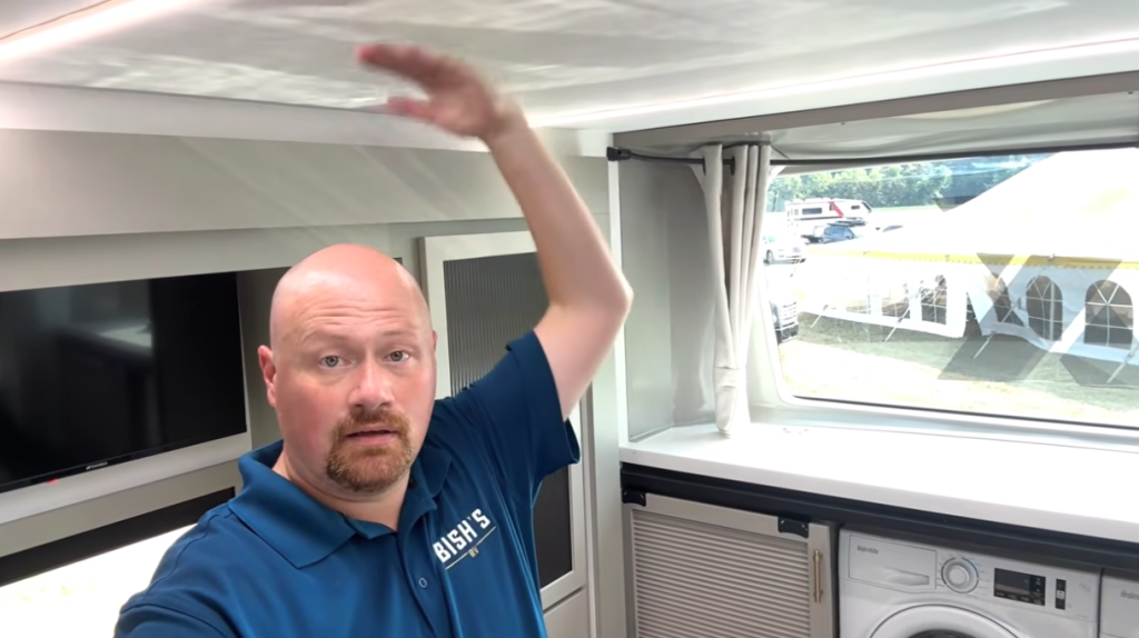 RV expert demonstrating ceiling height inside the 2026 Axiom Vendetta 4250 fifth wheel bedroom, showcasing the spacious front cap design with panoramic window and built-in washer-dryer units below.