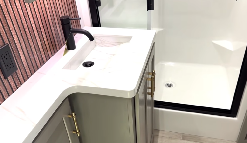 Bathroom vanity in the 2026 Axiom Vendetta 4250 fifth wheel featuring a white solid-surface countertop with black faucet, gold hardware cabinetry, wood-slat accent wall, and adjacent walk-in shower.