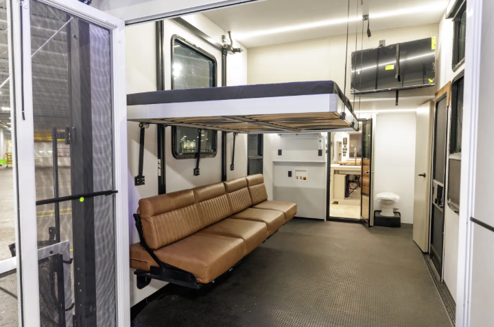 Garage of the 2026 Axiom Vendetta 4250 fifth wheel toy hauler featuring a Happy-Jack power lift bed system, fold-down leather sofa, rubberized flooring, and access to the half bath and main living area.