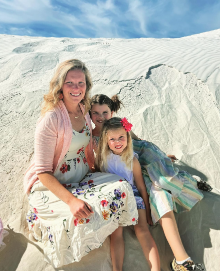 Woman and two young girls sitting together on bright white sand dunes under a blue sky, wearing colorful dresses and smiling for a cheerful family photo in a desert landscape.