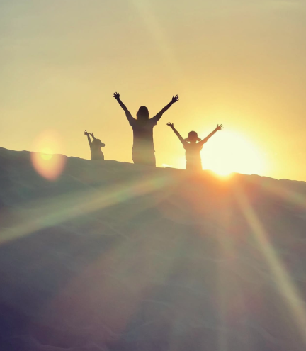 Silhouettes of three people standing on a sunlit sand dune at sunset with their arms raised, surrounded by warm golden light and dramatic lens flare.