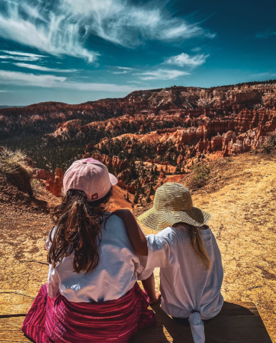 Two children sitting together at a scenic overlook, wearing sun hats and looking out over the dramatic red rock hoodoos and canyons of Bryce Canyon National Park under a bright blue sky.