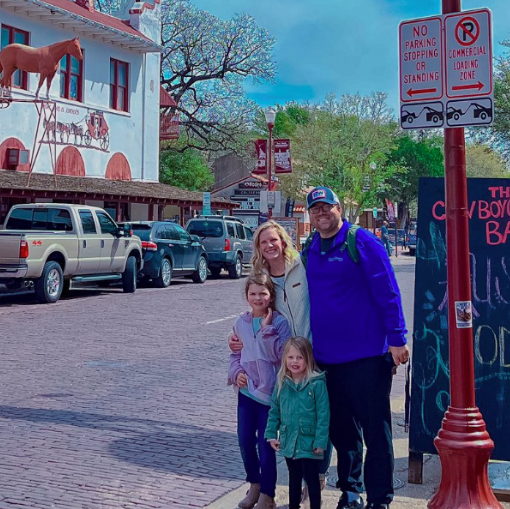 Family standing together on a historic brick street in a western-themed district, surrounded by parked trucks, rustic storefronts, and signage, capturing a cheerful sightseeing moment.