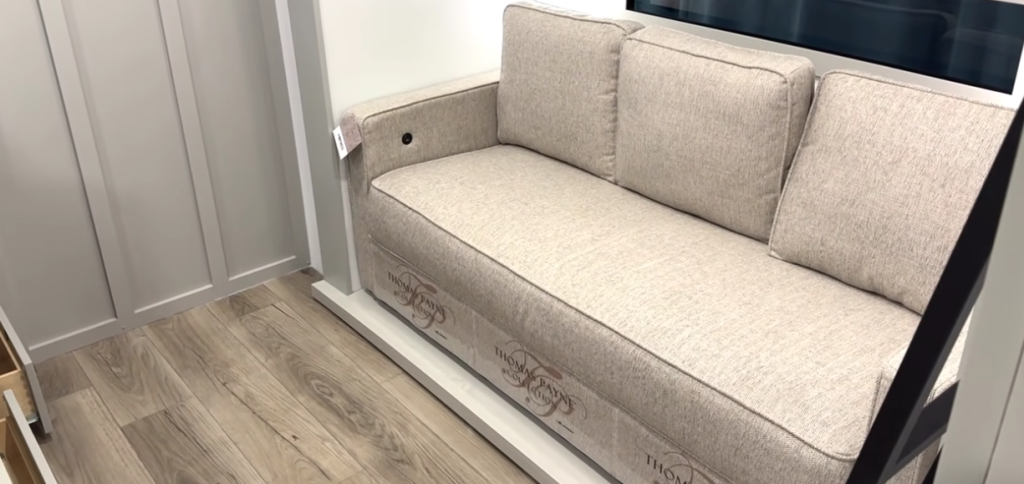 RV rear bunk room sofa with light gray upholstery, three plush cushions, and built-in armrest storage, shown on wood-look flooring with modern wall paneling