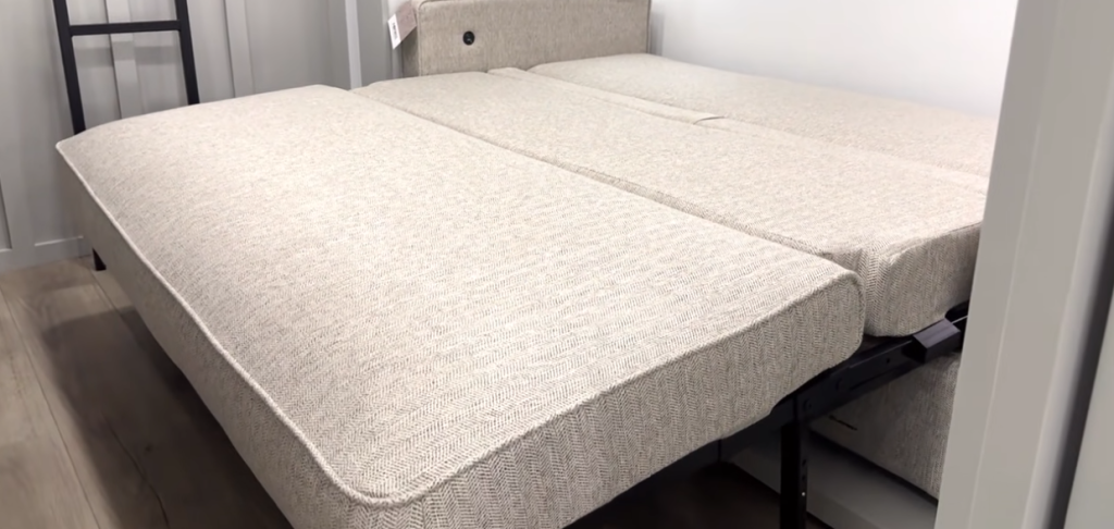 RV bunk room tri-fold sofa fully extended into a bed, featuring light gray textured cushions and metal support legs on wood-look flooring
