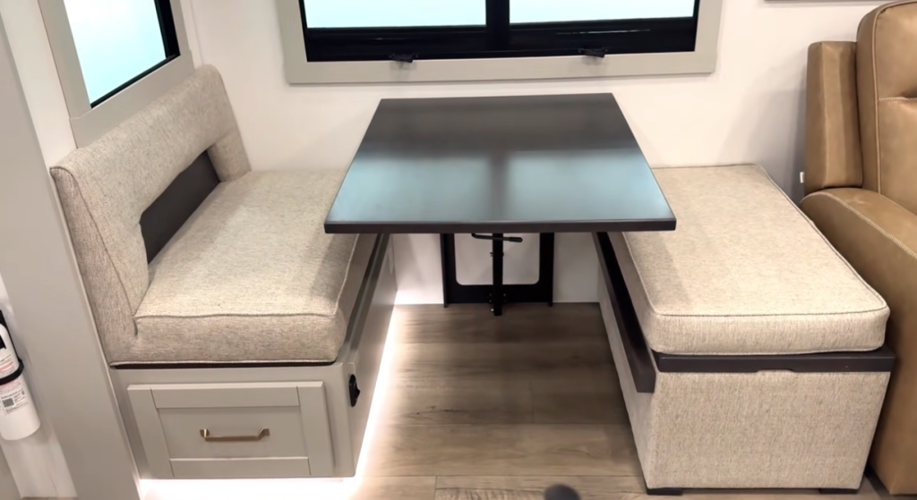 RV dinette with dual bench seating, storage drawers, and a dark wood table inside a modern travel trailer interior with neutral upholstery and under-seat accent lighting.