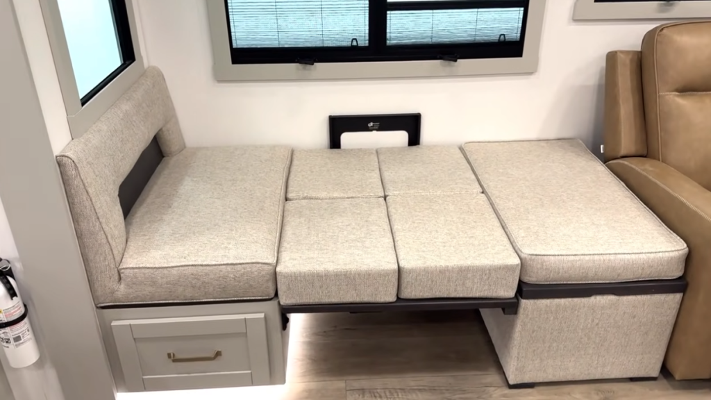 RV dinette converted into a sleeper bed with extended cushions, light gray upholstery, built-in storage drawer, and modern interior design