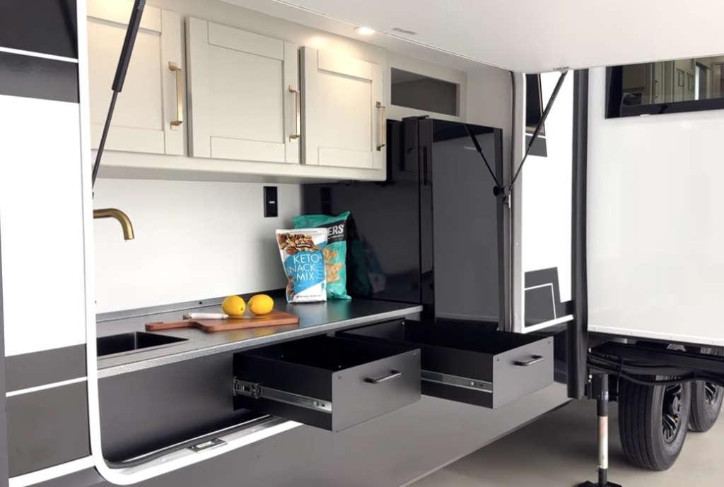 Exterior camp kitchen on the 2026 Brinkley Model Z Air 315 featuring upper shaker cabinets, a brass faucet, stainless countertop, 12V outdoor refrigerator, and dual pull-out storage drawers with snacks and cutting board displayed.