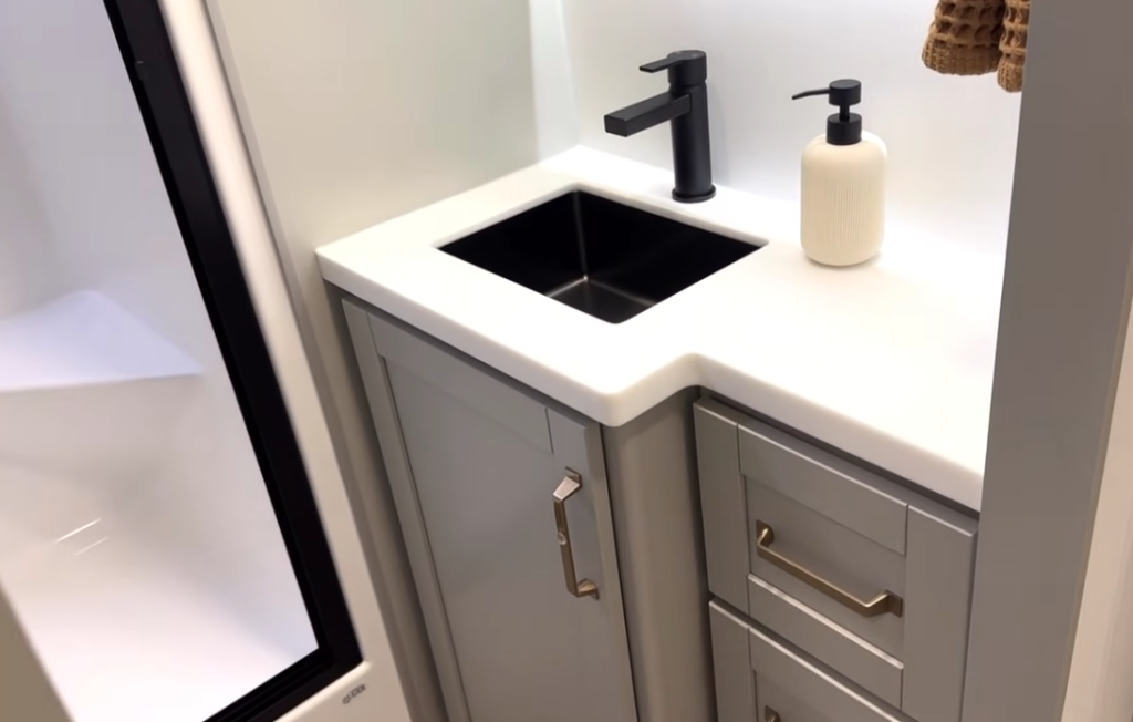 RV bathroom vanity in the 2026 Brinkley Model Z Air featuring a solid-surface countertop, black modern faucet, stainless sink, gray shaker cabinetry, and part of the one-piece fiberglass shower.
